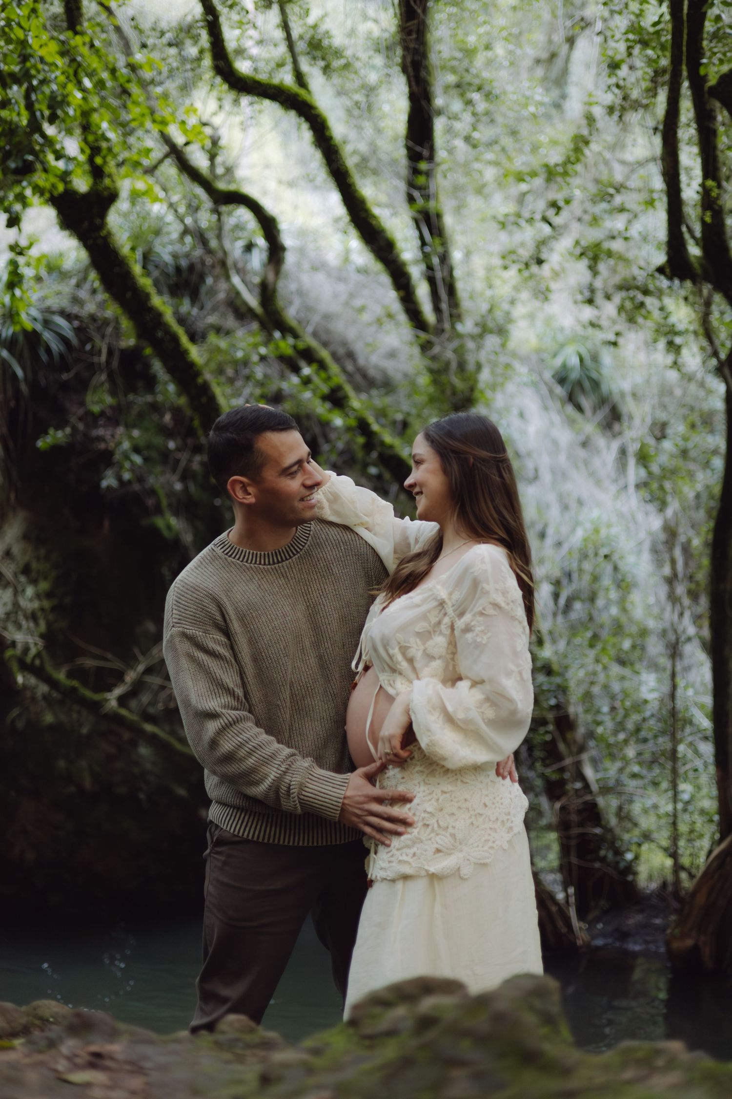 Couple sharing a tender moment in a lush forest, man holds pregnant woman's belly near a waterfall.