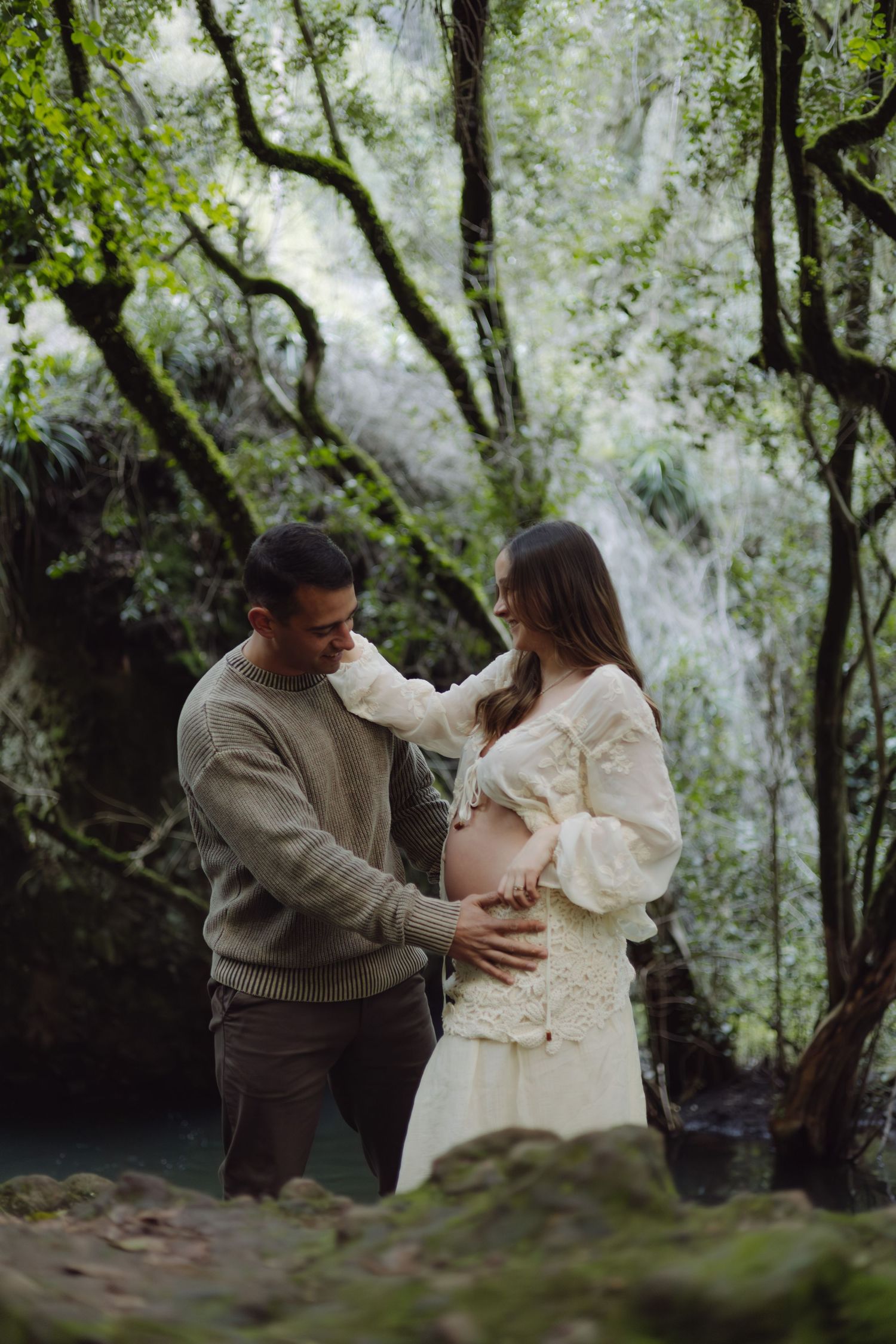 Expectant couple standing together in green woodland, man gently touches pregnant partner's baby bump.