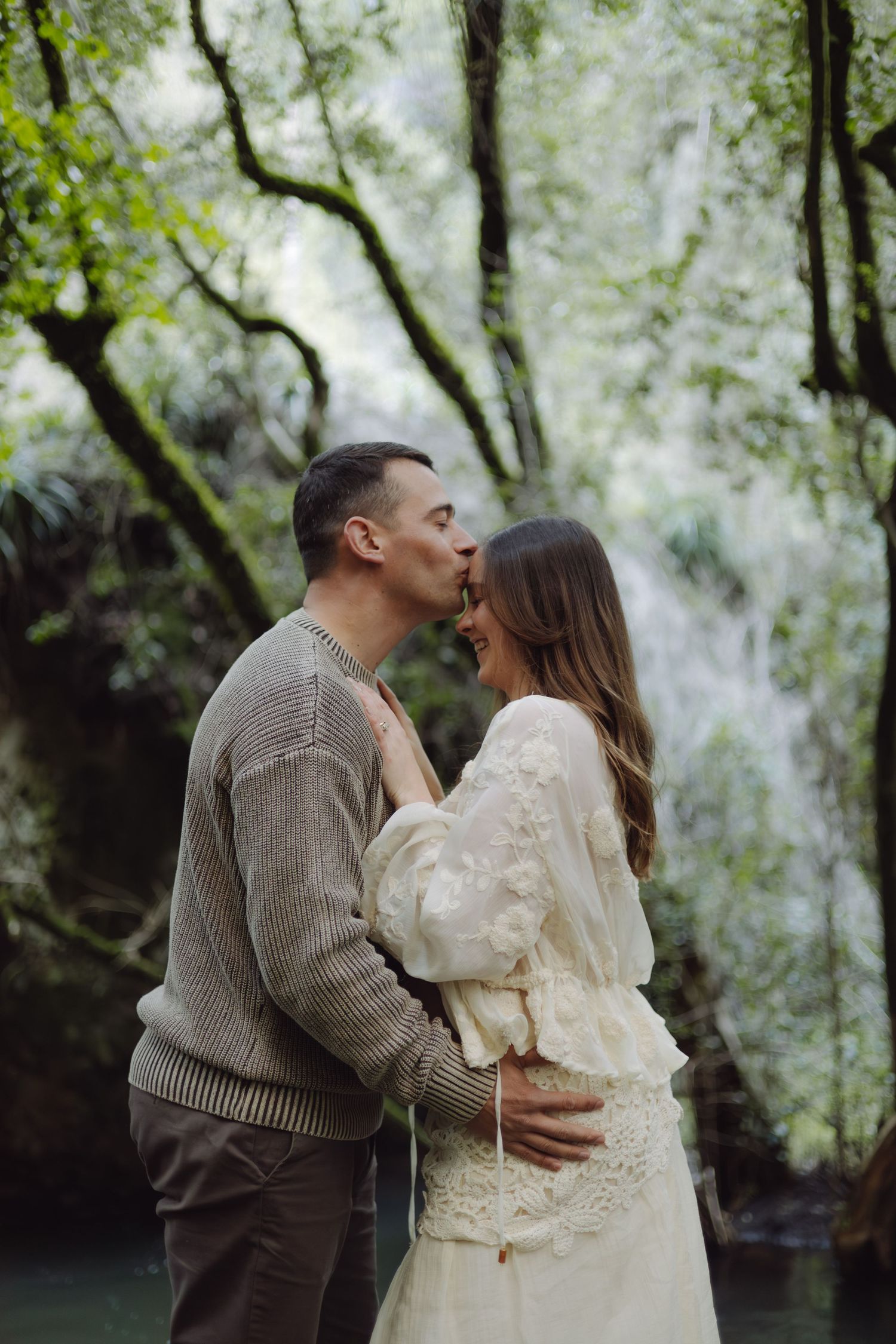 A couple shares a romantic kiss in front of a lush waterfall, man in knit sweater embracing woman in white lace dress.