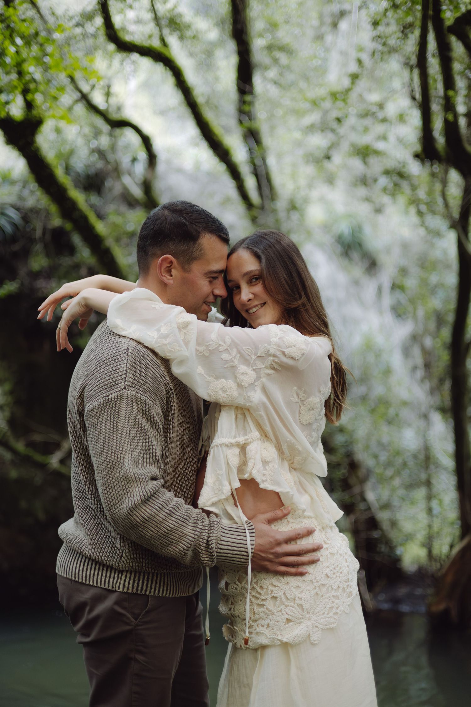 Couple embracing tenderly in front of a lush waterfall, woman in white lace dress and man in brown knit sweater.