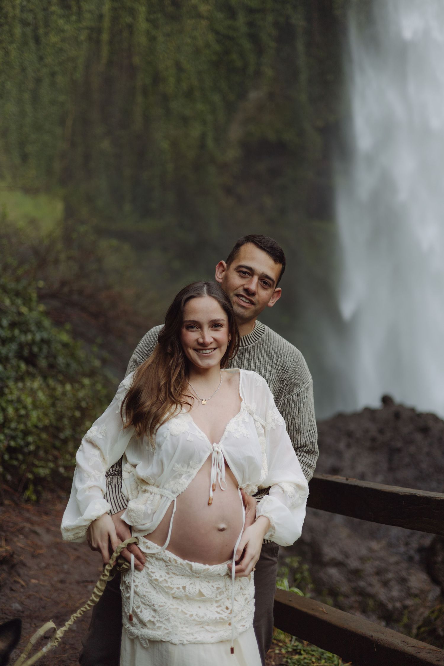 Smiling pregnant couple posing in front of a stunning waterfall, woman in white boho maternity outfit.