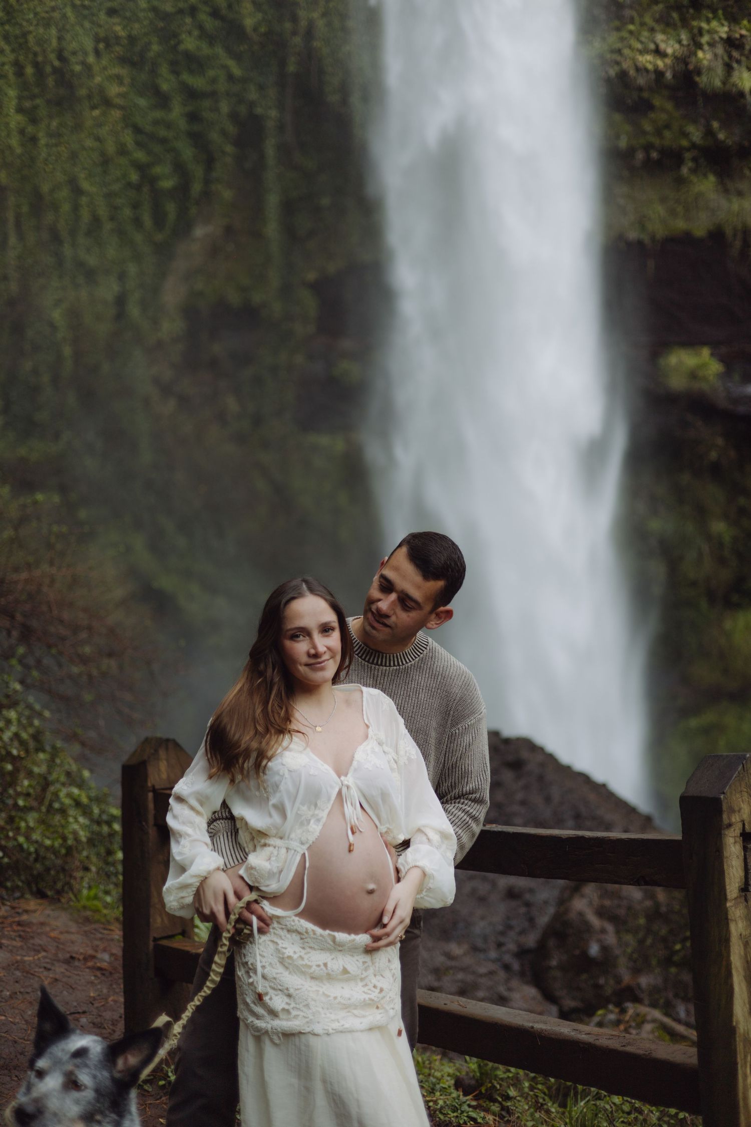 Expecting couple with dog posing before dramatic waterfall, woman in white dress showing baby bump, man in gray sweater embracing her.