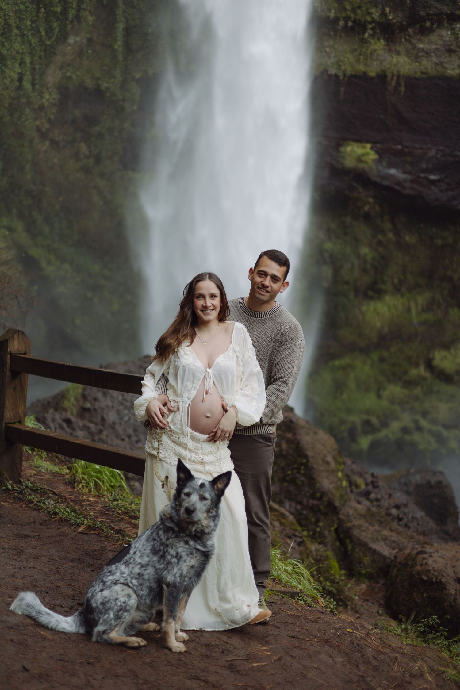Happy expecting couple with their blue heeler dog posing before a majestic waterfall during maternity shoot.