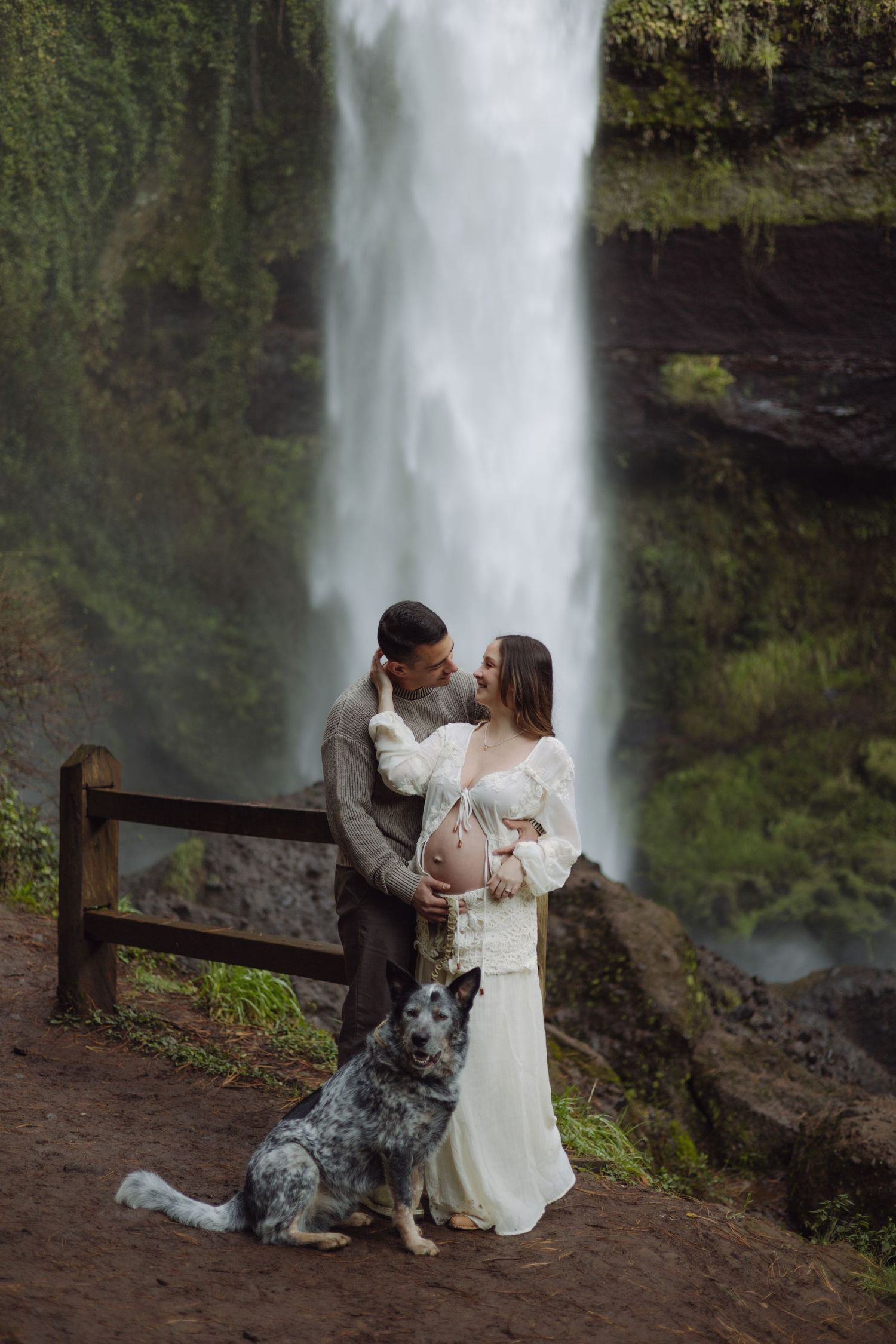 Pregnant couple kissing before tall waterfall, blue heeler dog sitting beside them, woman in white lace dress with exposed belly.