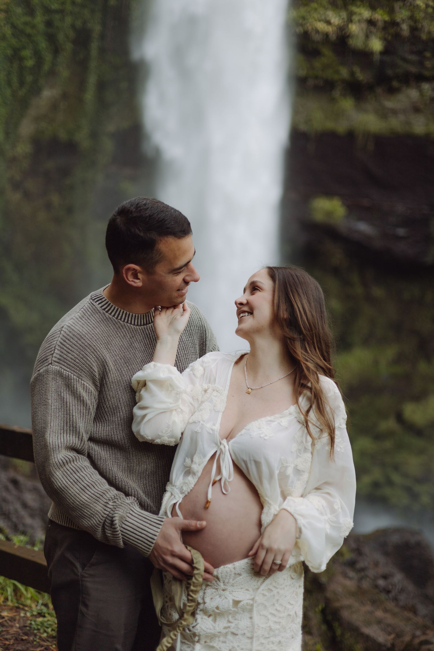 Couple smiling at each other in front of a waterfall, woman in white lace dress showing pregnant belly.