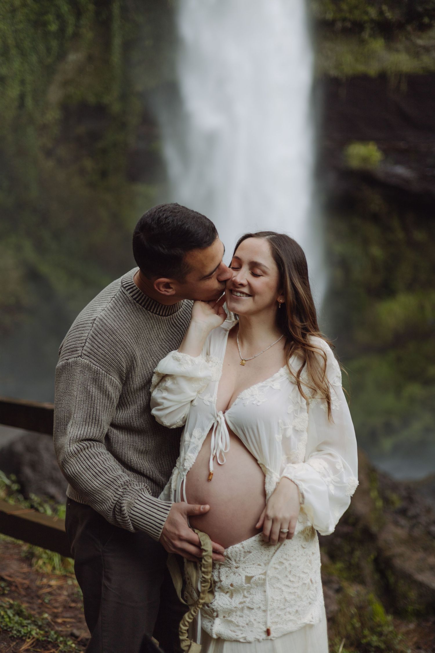 Man kissing pregnant woman's cheek in front of a waterfall, both cradling her baby bump tenderly.