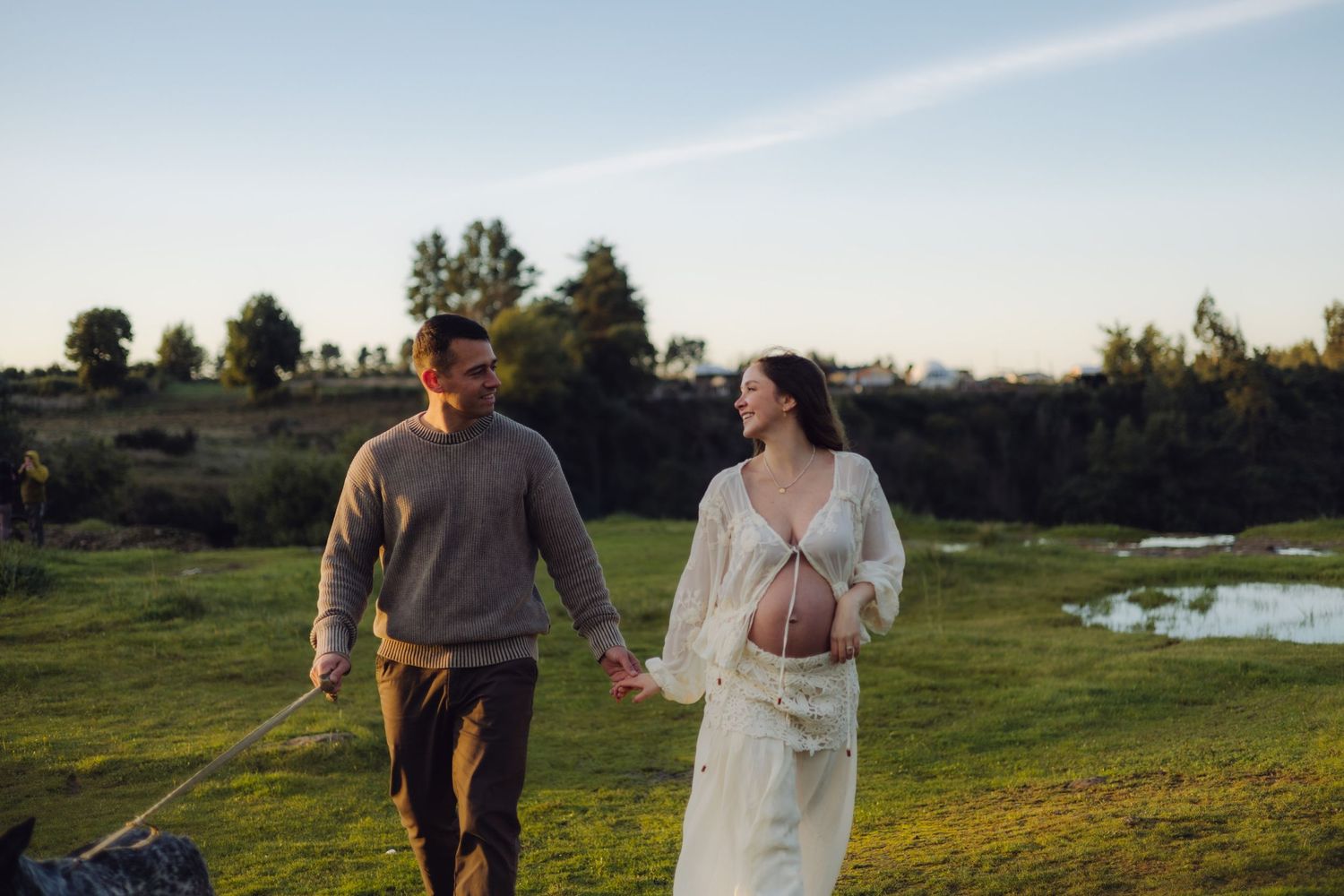 Pregnant couple holding hands walking in green field at golden hour, woman in white dress showing baby bump.