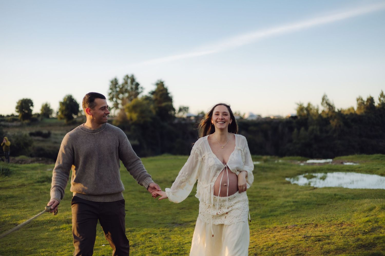 Smiling pregnant woman in white dress holds hands with man in gray sweater walking through green field at sunset.