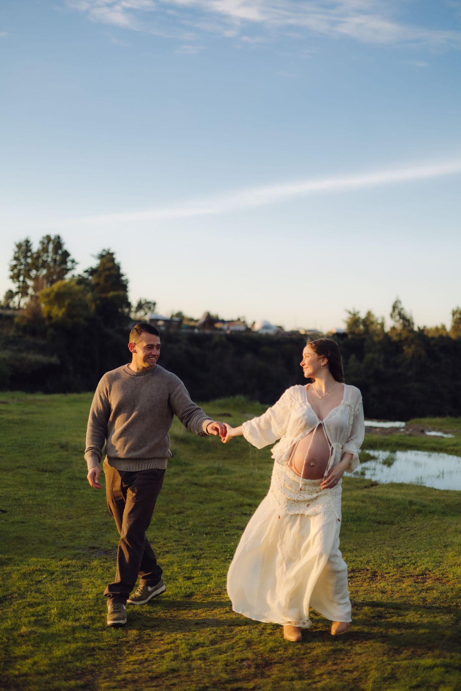 Pregnant woman in white dress holds hands with man in gray sweater walking on green field at sunset.
