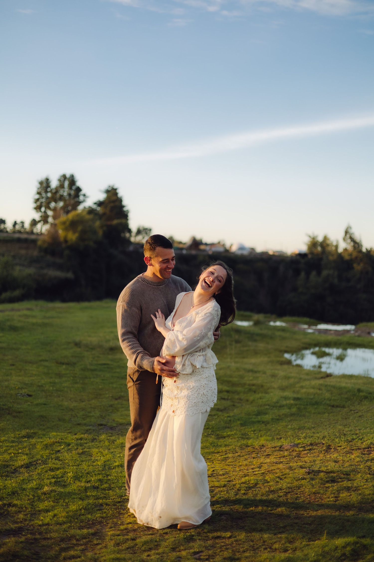 Happy couple embracing outdoors at golden hour, bride in white gown laughing joyfully with groom in gray sweater.