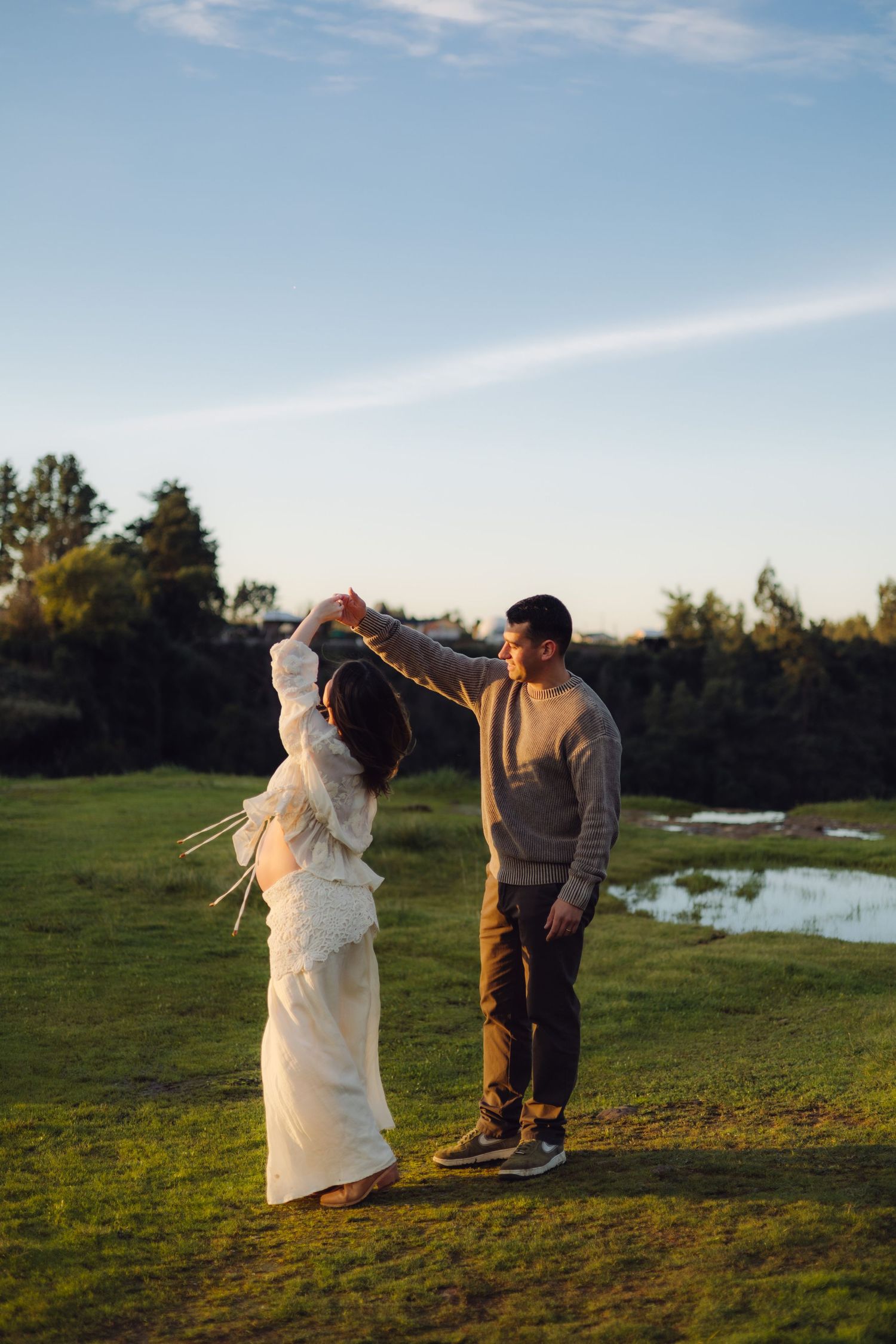 Couple dancing outdoors on green meadow at sunset, man spinning woman in white dress.