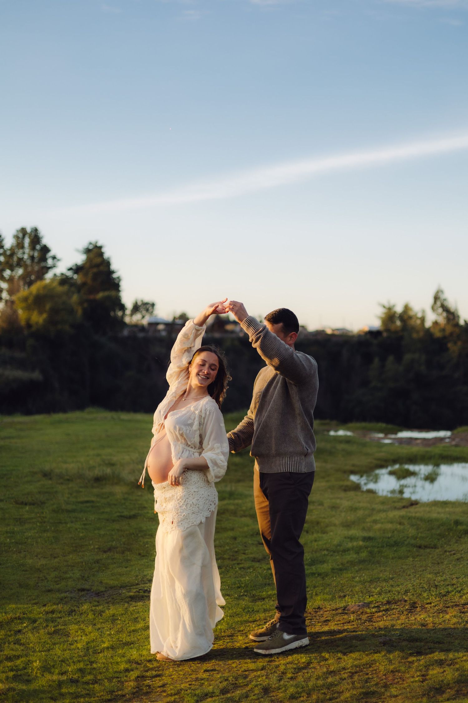 Couple dancing outdoors at golden hour, pregnant woman in white lace dress twirling with partner on green grass.