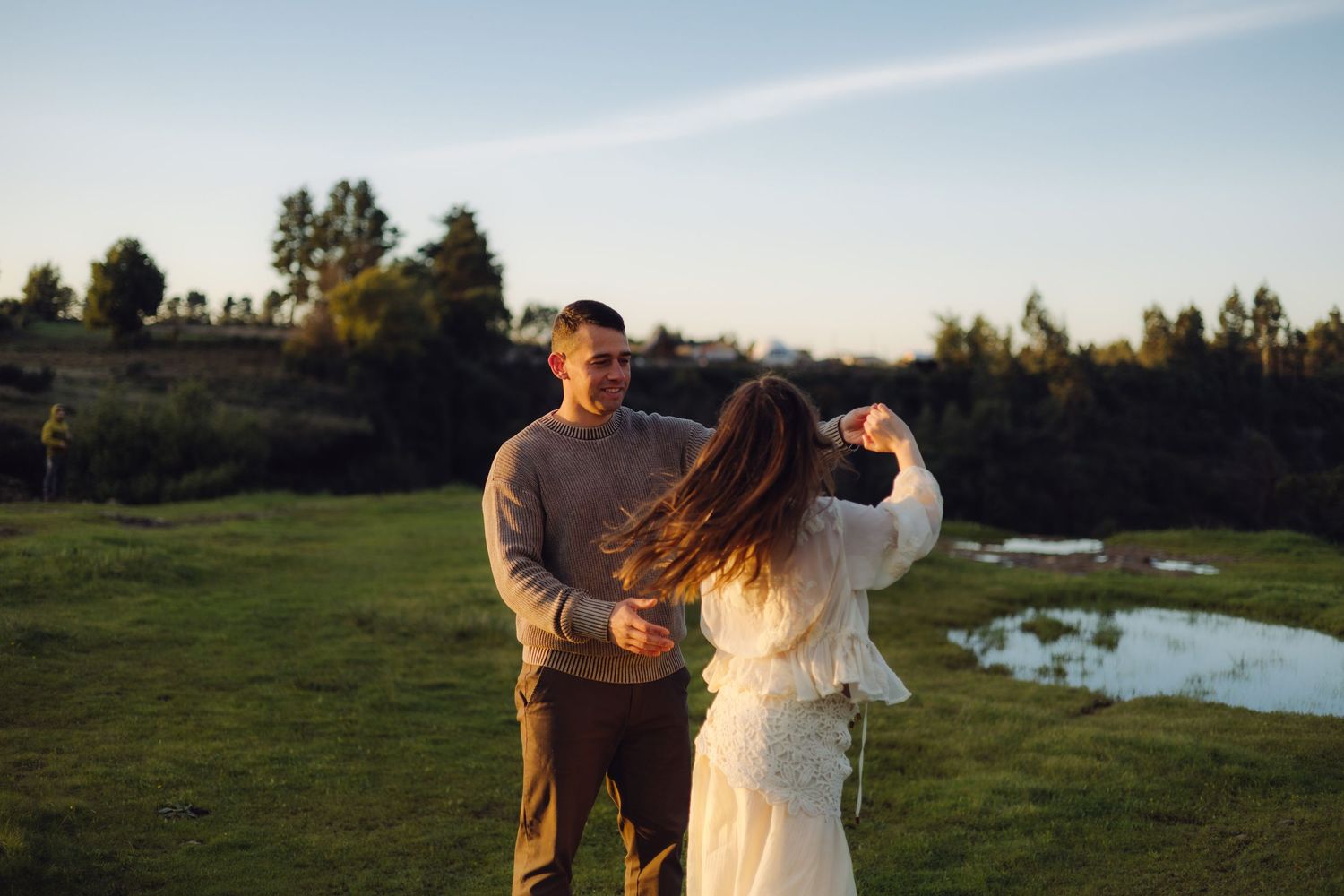 Couple dancing together in a sunlit green field at golden hour near a small pond.