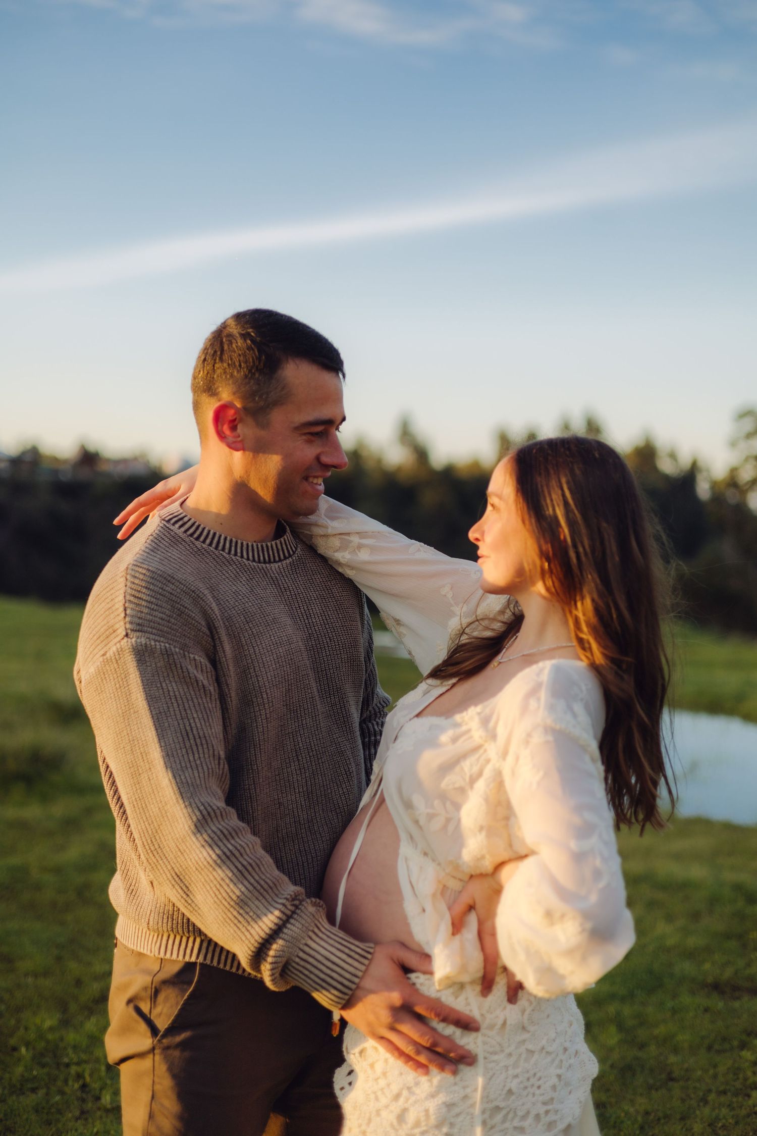 Smiling couple outdoors at golden hour, pregnant woman in white lace dress embracing man in gray knit sweater in green field.