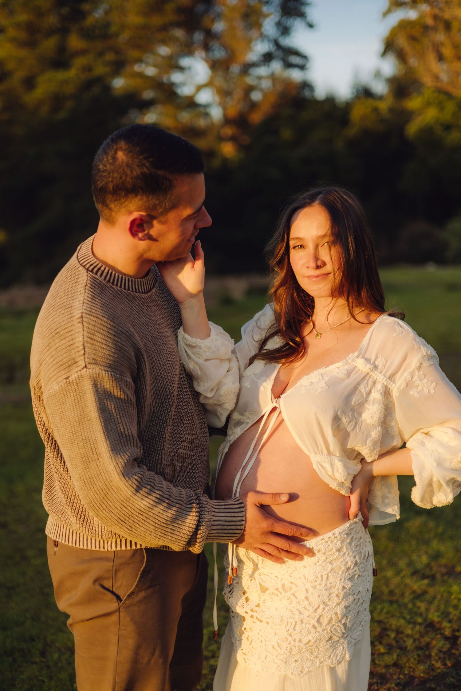 Expectant couple sharing tender moment at sunset, woman in white dress cradling baby bump while partner holds her face gently.