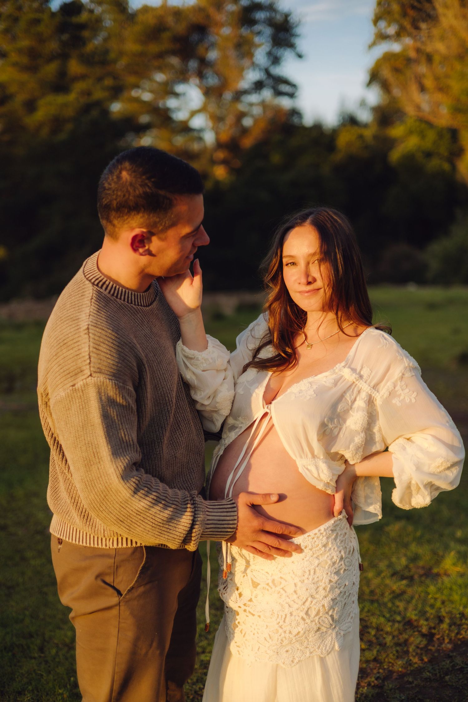 Couple in golden hour light outdoors, pregnant woman in white dress with partner in tan sweater cradling her baby bump.