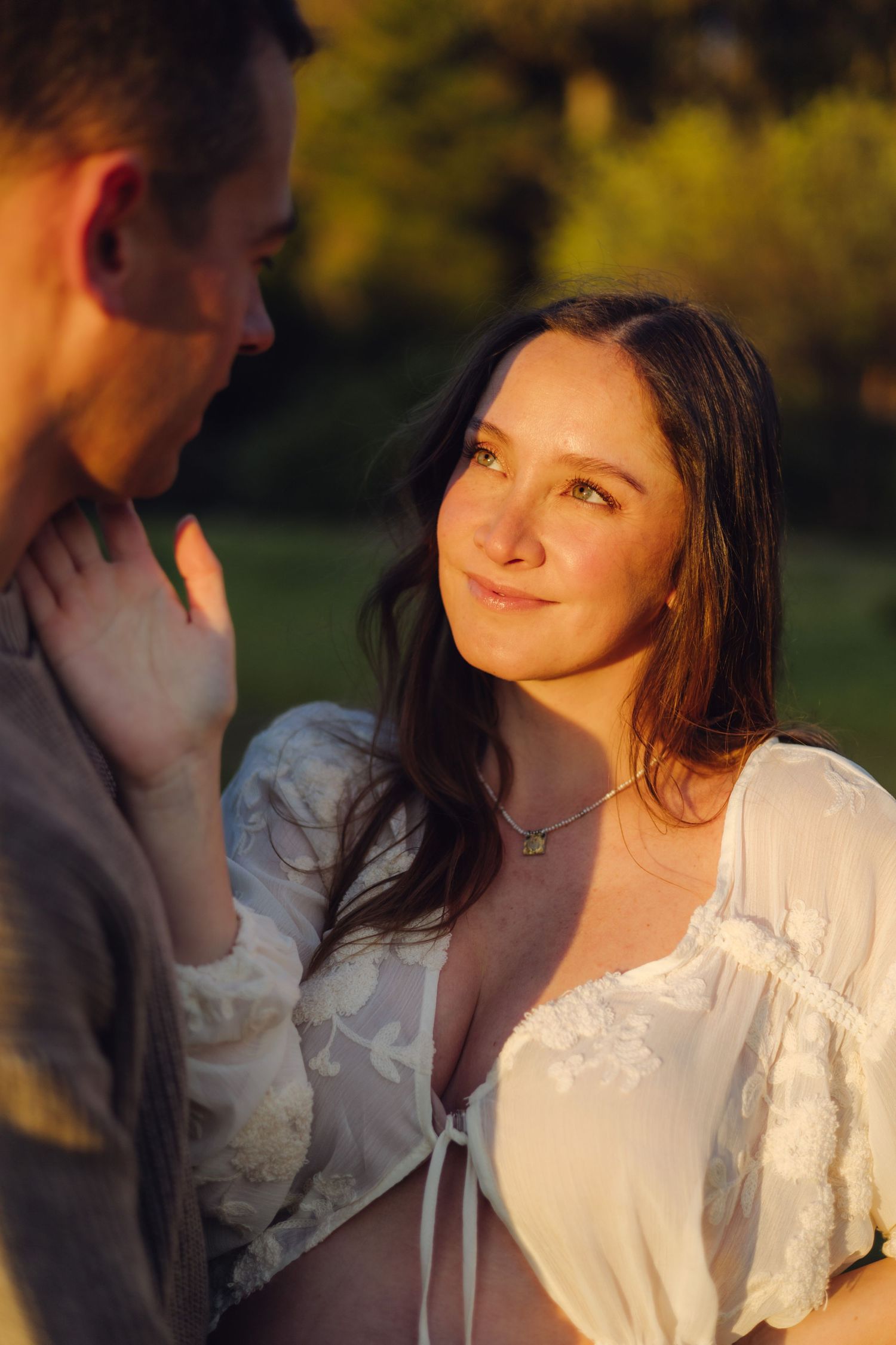 A woman in a white lace top smiles warmly at a man during a golden hour outdoor couples portrait.