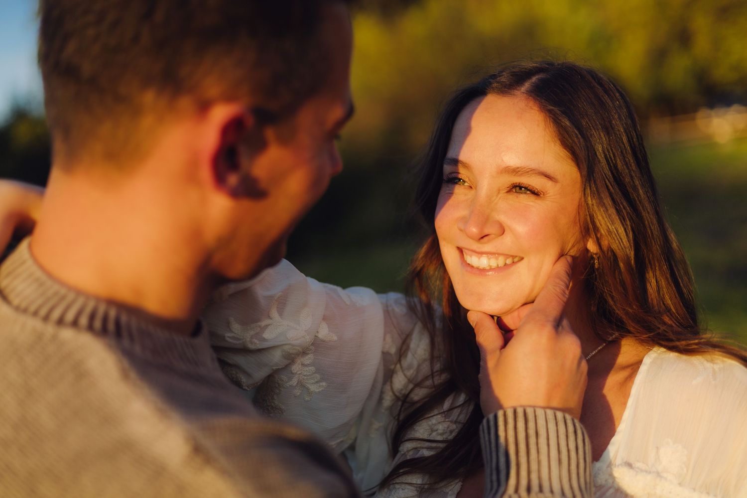 A smiling young woman gazes at a man outdoors in warm golden sunlight during a romantic moment.