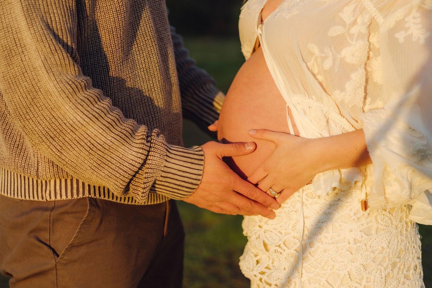 Close-up of couple holding hands over pregnant belly in golden light, woman wearing white lace dress.