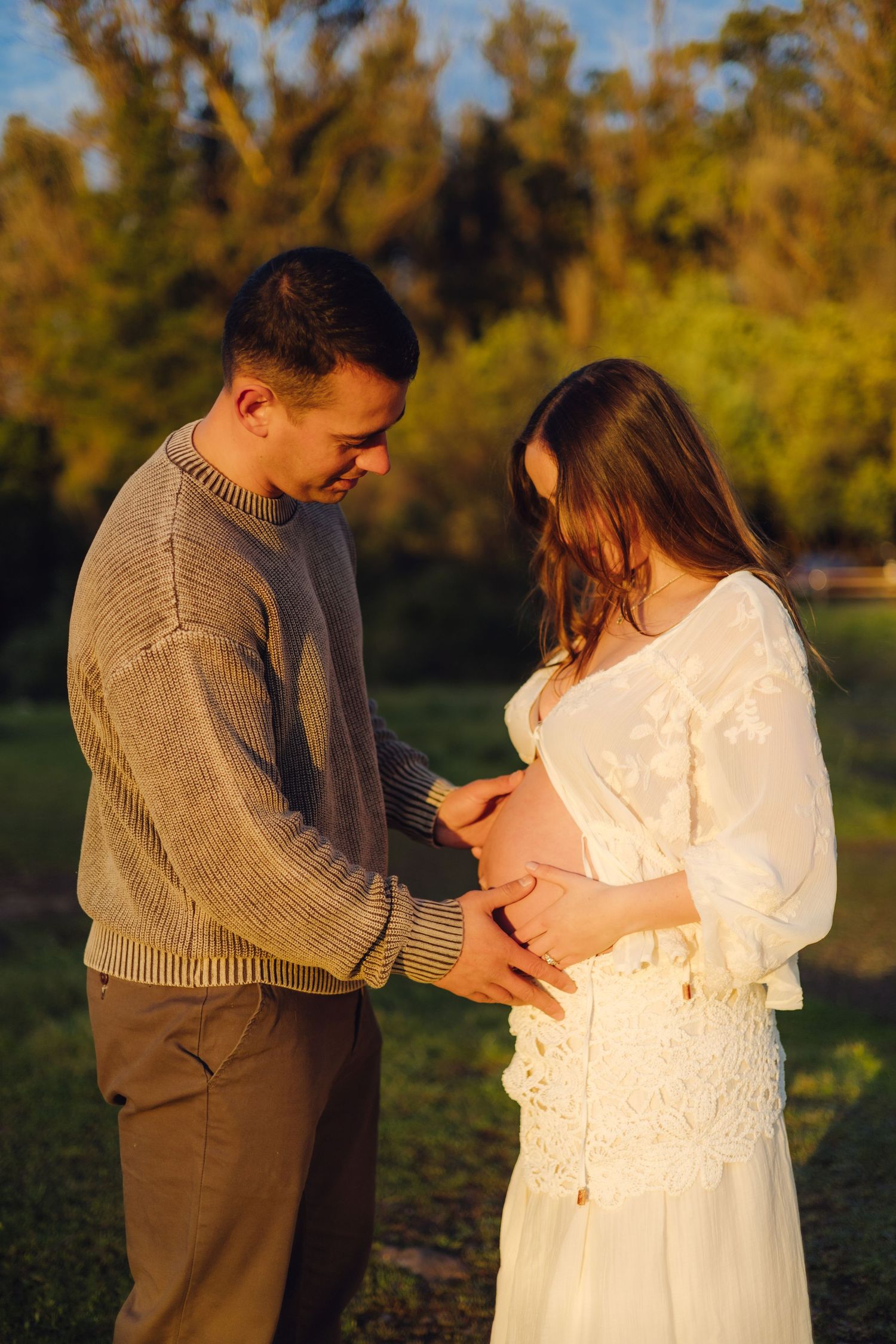 Couple tenderly cradling pregnant belly outdoors at golden hour, man in beige sweater and woman in white lace dress.