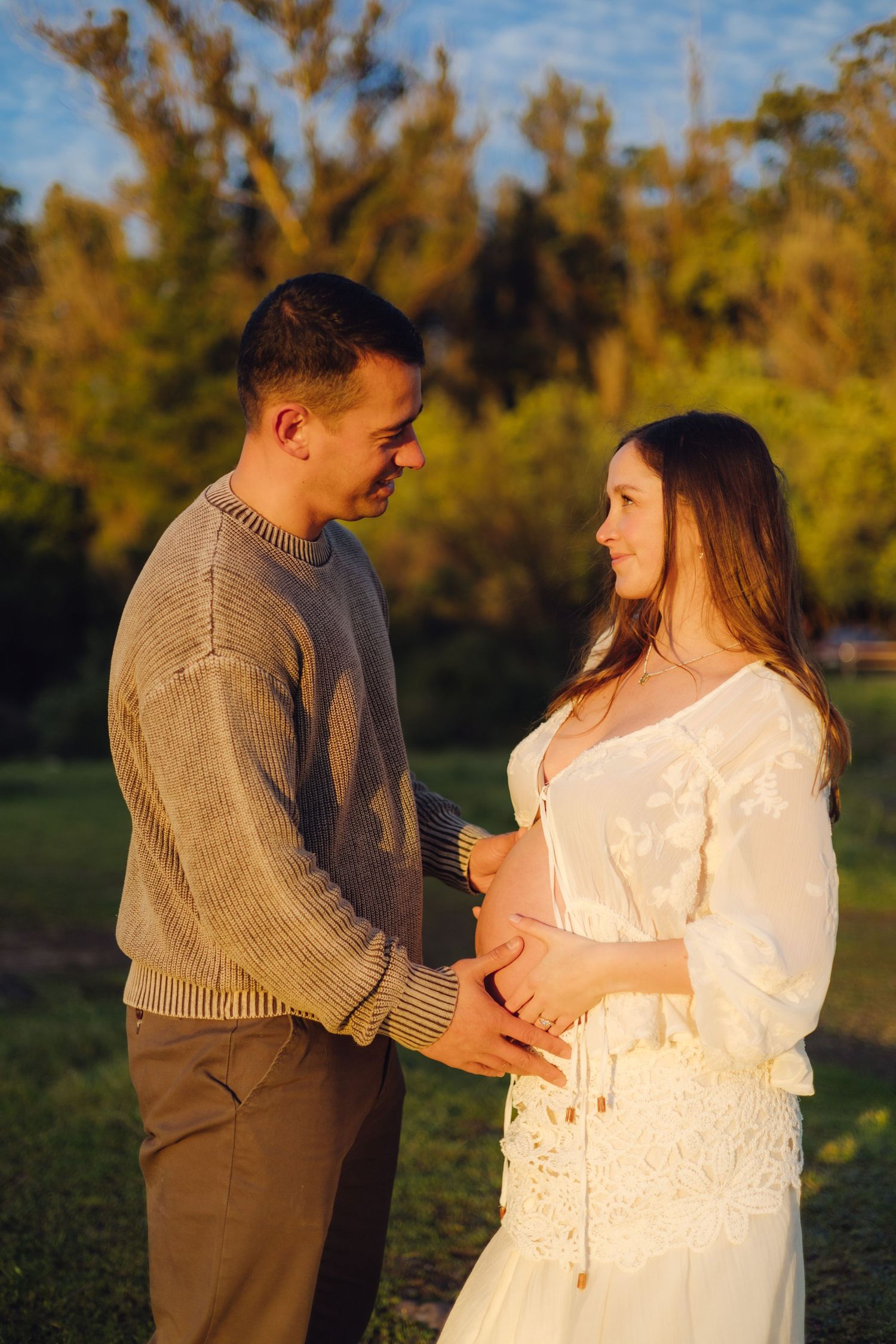 Couple tenderly cradling pregnant belly at golden hour in park, man in gray sweater and woman in white lace dress.