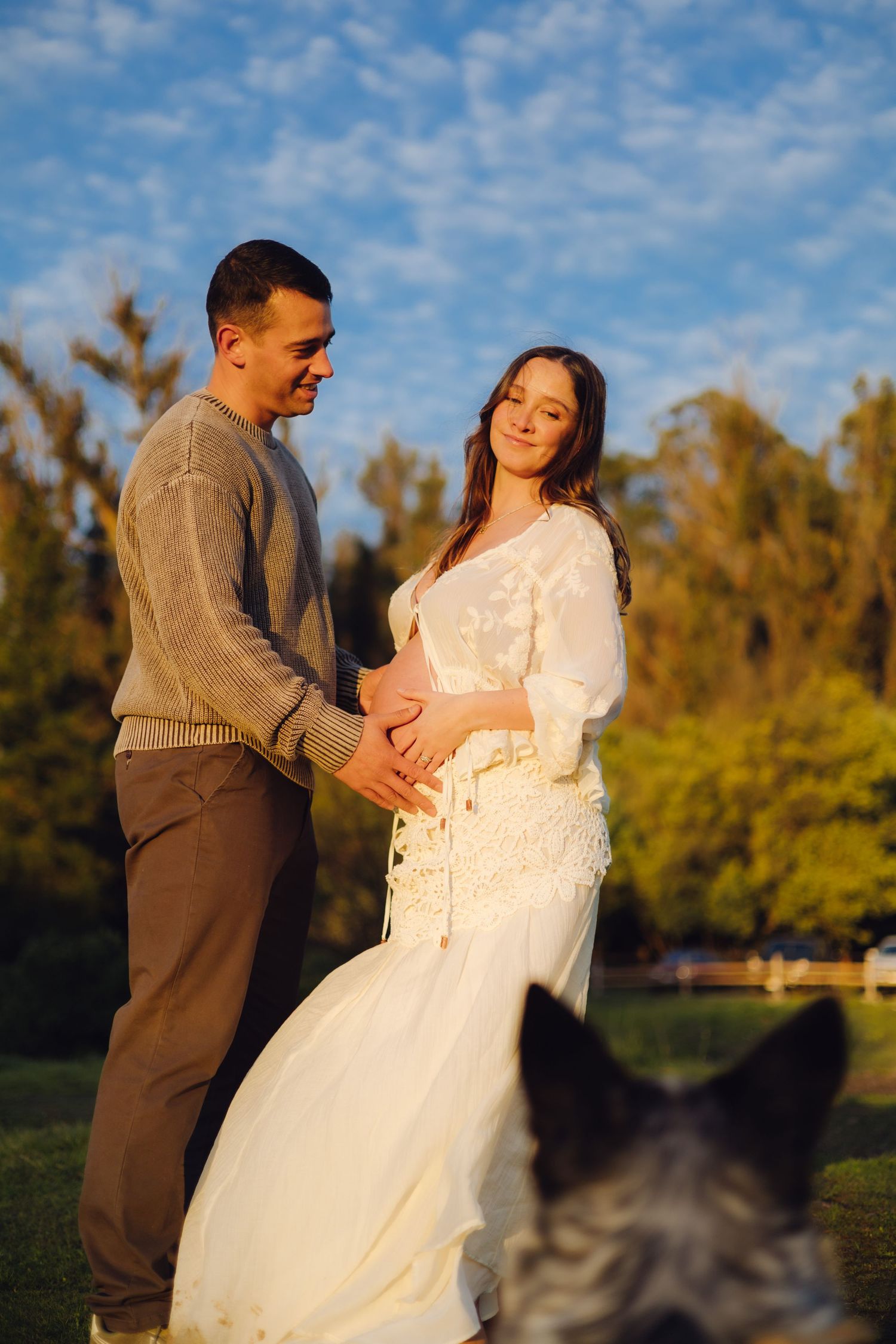 Couple tenderly holding pregnant belly outdoors at golden hour, with a dog in foreground and blue sky behind.