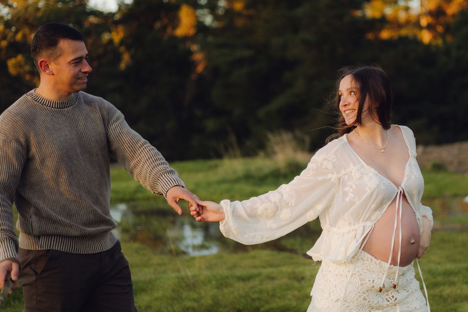 Happy couple holding hands in autumn park, pregnant woman in white dress laughing with partner at golden hour.