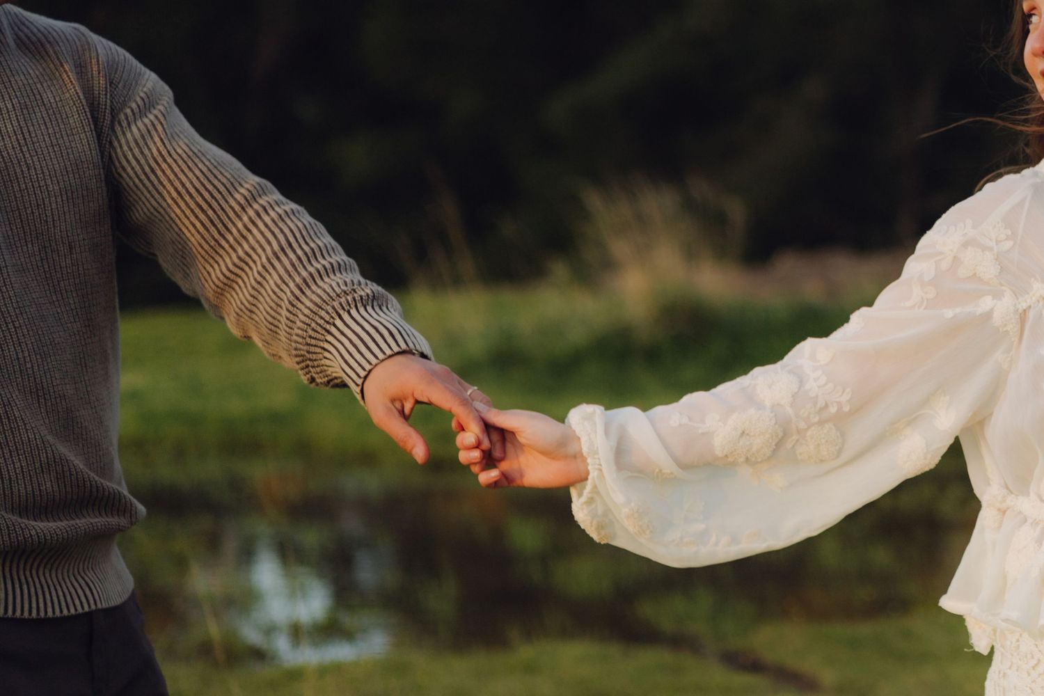 Close-up of couple holding hands outdoors, woman in white lace dress and man in gray knit sweater near a peaceful pond.