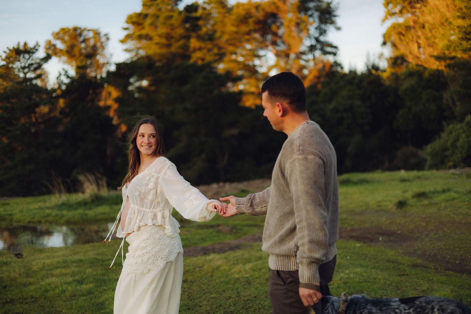 Smiling pregnant woman in white lace dress holds hands with man in gray sweater during golden hour outdoor photoshoot.