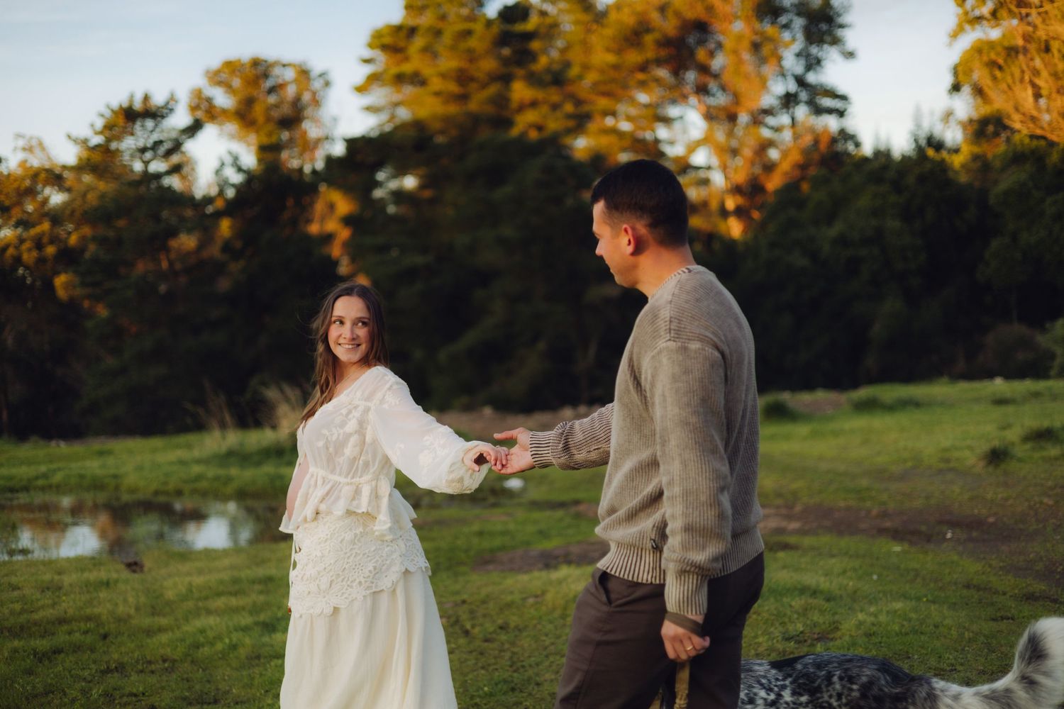 Pregnant woman in white lace dress smiling while holding hands with partner in gray sweater during golden hour outdoors.