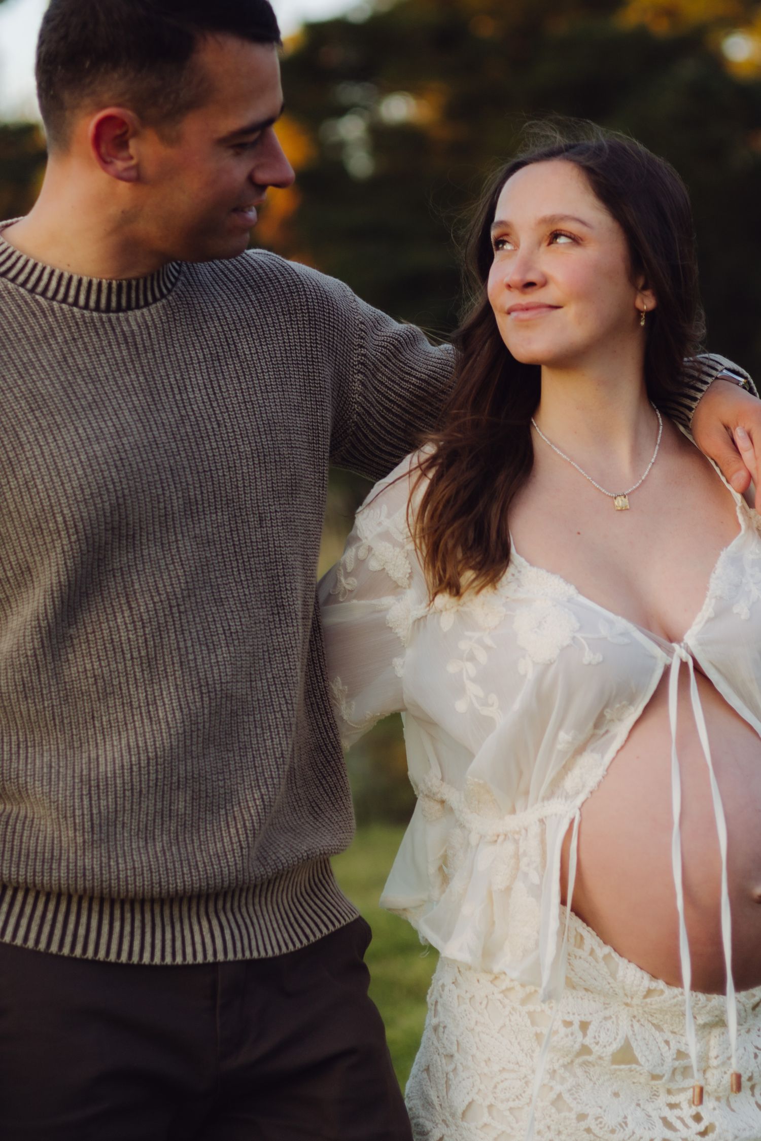 A couple shares a tender moment outdoors, the woman in a white lace maternity gown, the man in a gray sweater.