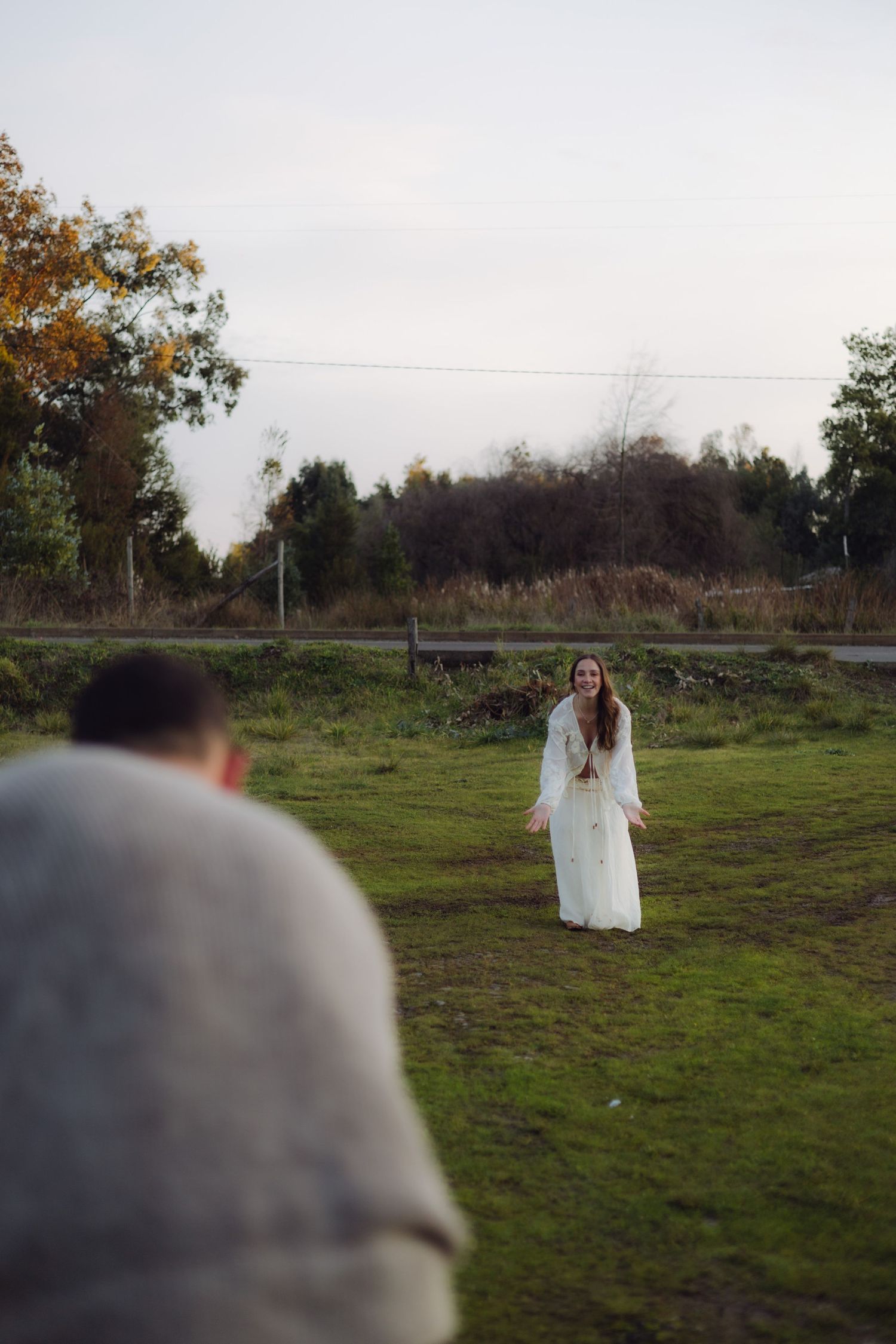 A bride in a white dress smiles at a man in a gray sweater in a green field with autumn trees in the background.