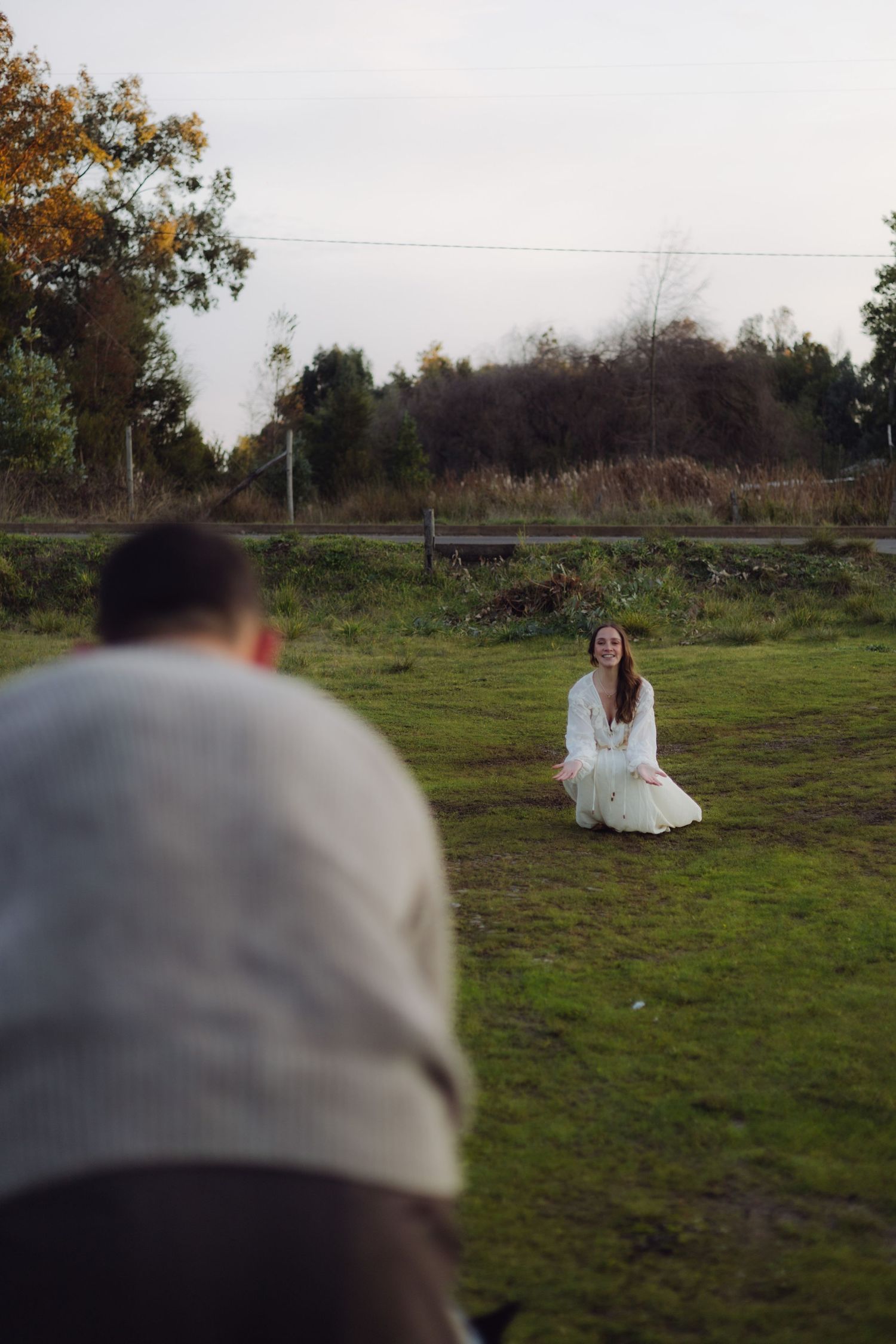 Photographer capturing smiling woman in white dress kneeling on grass in an outdoor field setting.