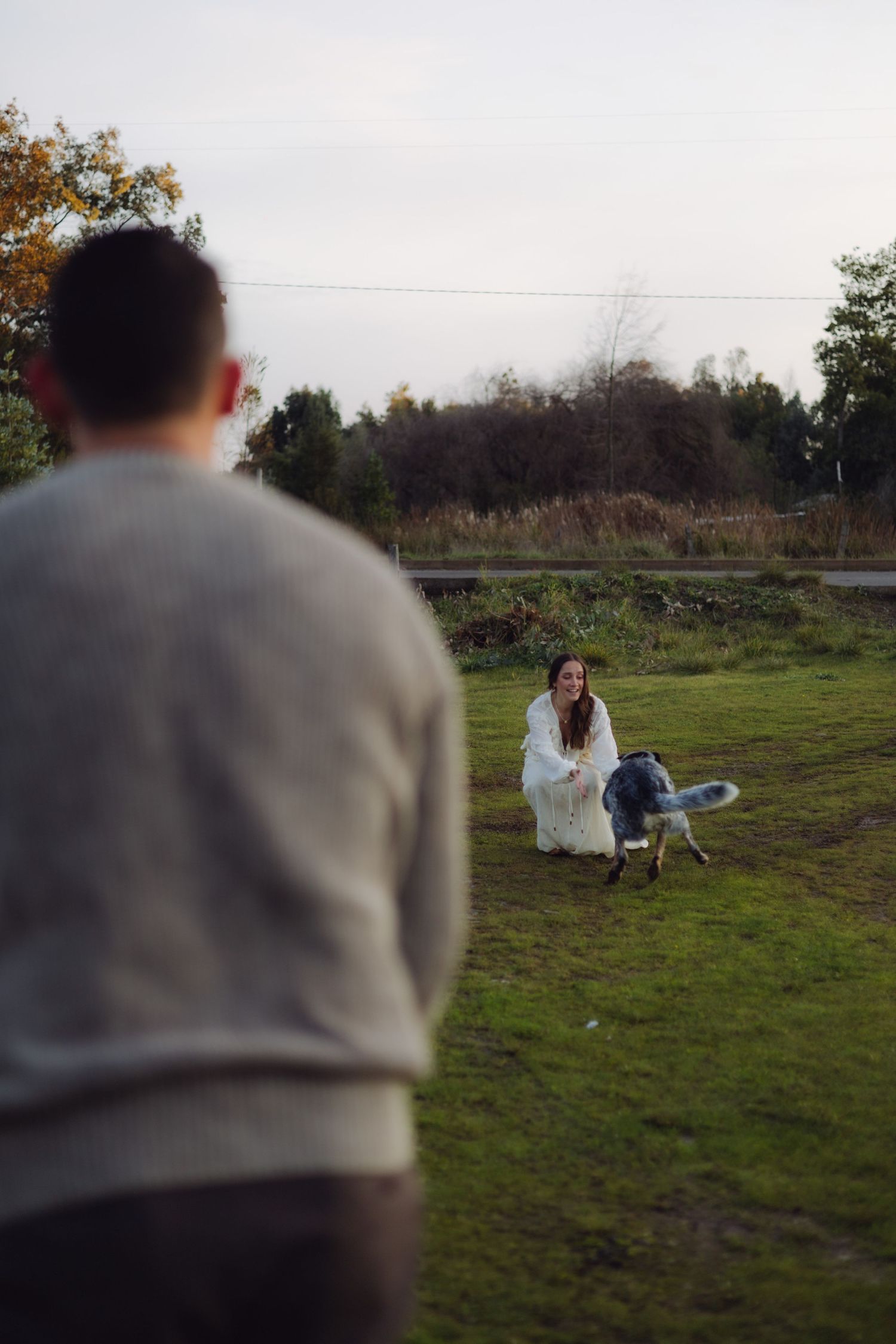 Pregnant woman in white dress crouching on grass with dog while man watches in foreground.