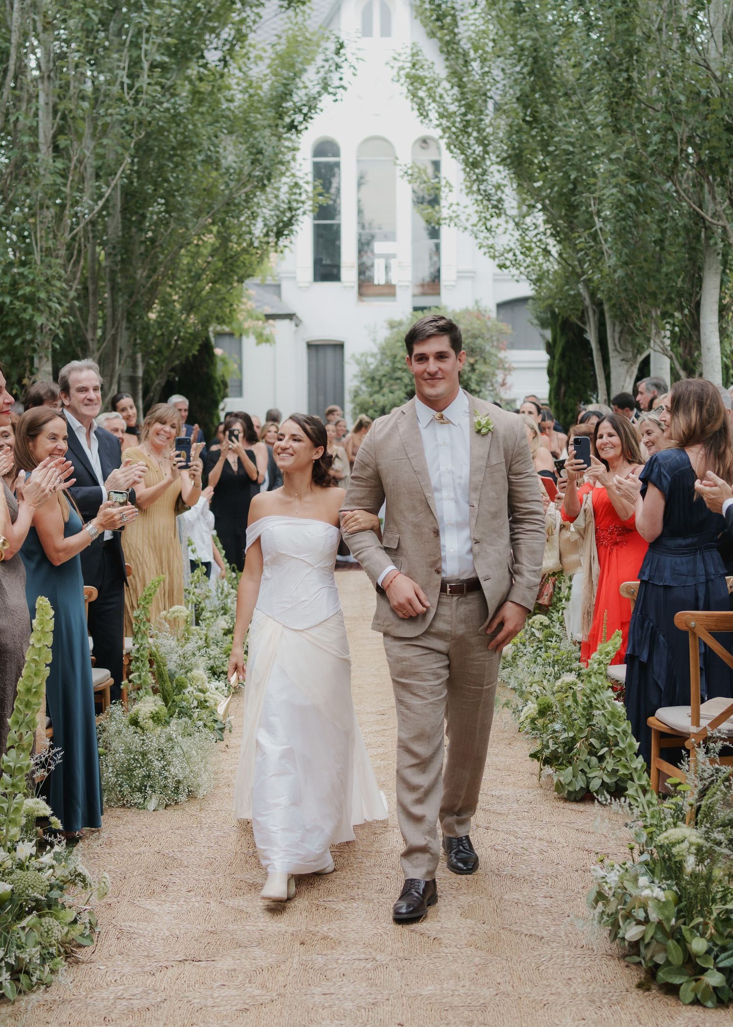 Bride and groom walking down outdoor aisle lined with trees and guests applauding at garden wedding ceremony.