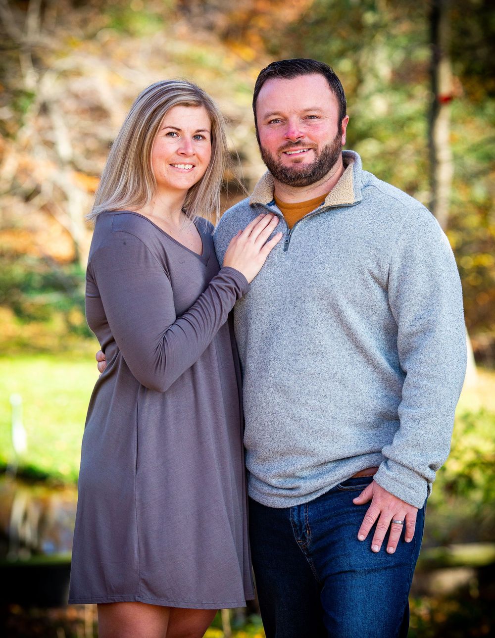 A smiling couple poses outdoors in fall foliage, woman in gray dress and man in light gray pullover and jeans.