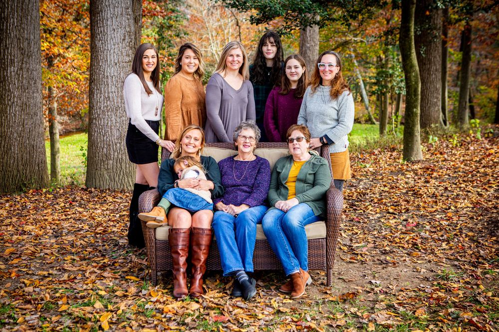 Multi-generational family portrait outdoors in autumn, posing on a bench surrounded by colorful fallen leaves and trees.