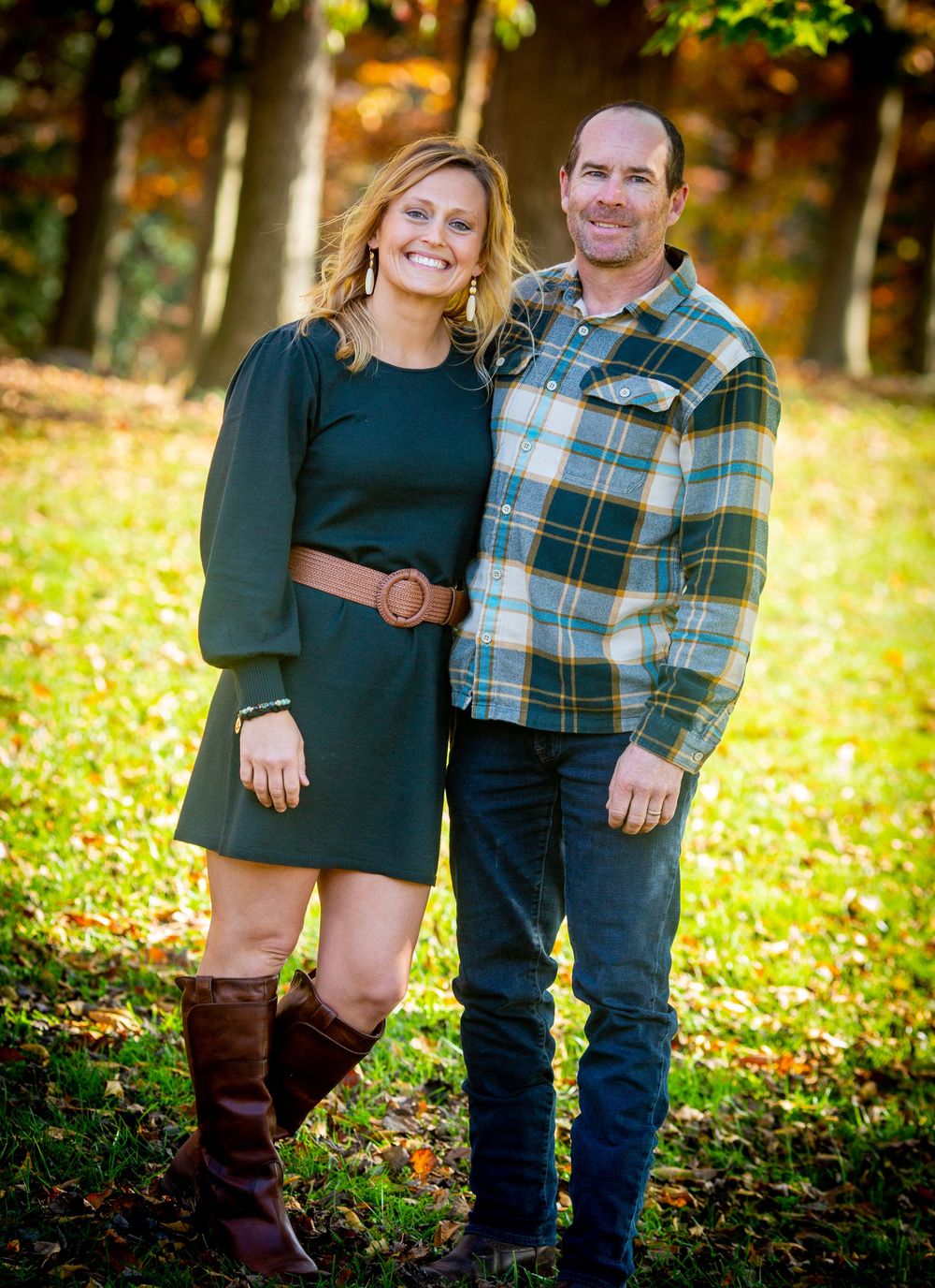 A smiling couple poses outdoors in autumn, woman in green dress with brown boots, man in plaid shirt, surrounded by fall foliage.
