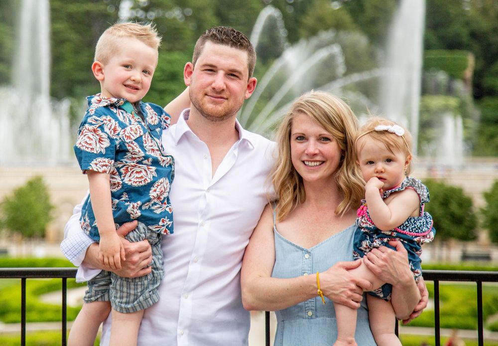 Happy family of four posing outdoors near a fountain, parents holding two young children on a sunny day.