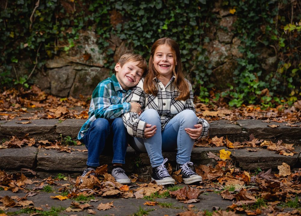Two smiling children in plaid shirts sit together on stone steps surrounded by colorful autumn leaves outdoors.