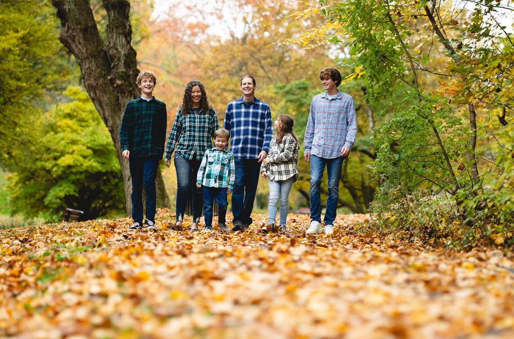 Family of six walking on a leaf-covered path surrounded by colorful autumn foliage in a park.