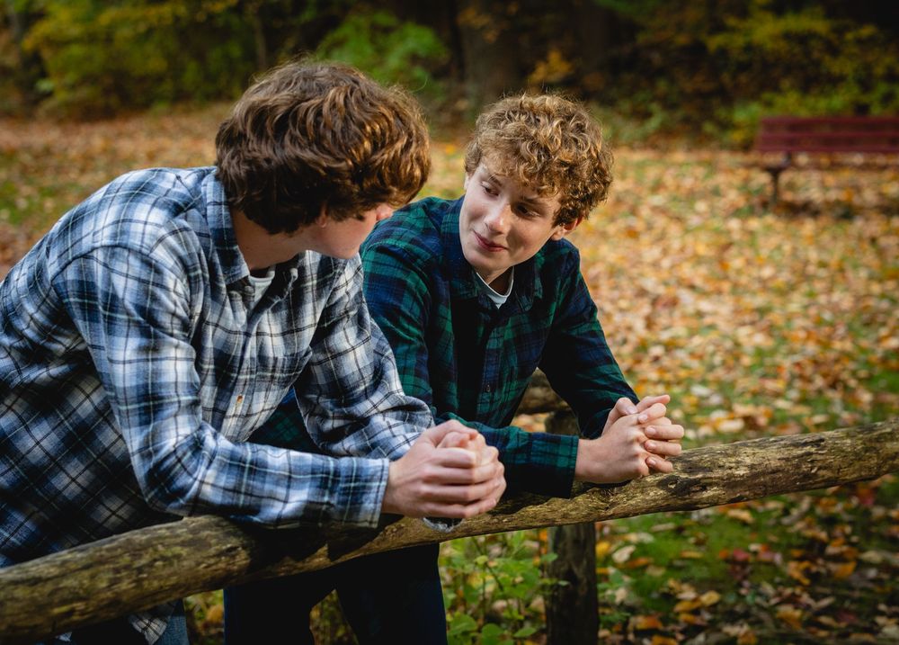 Two teenage boys in flannel shirts lean on a wooden fence, chatting outdoors among colorful autumn leaves.