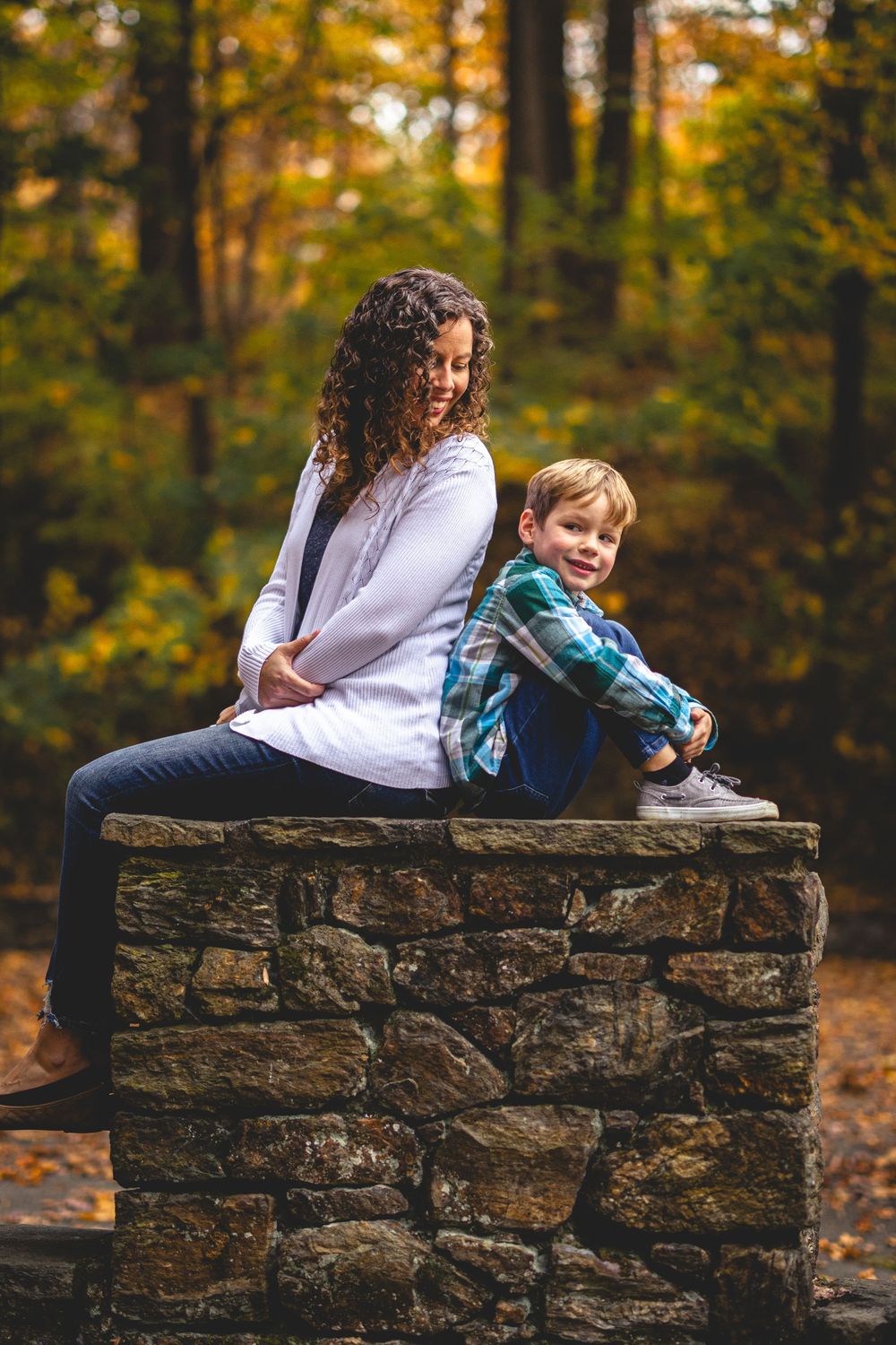A woman and young boy sit back-to-back on a stone wall surrounded by colorful autumn foliage.