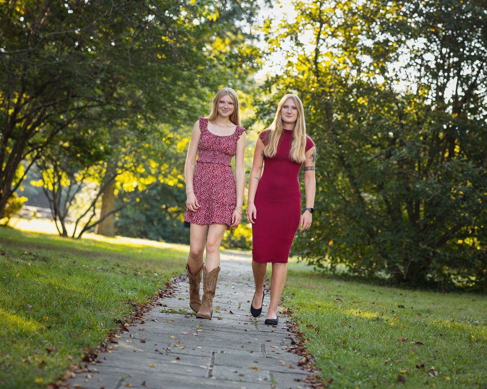 Two blonde women in elegant dresses walk along a brick path surrounded by lush green trees in a park setting.