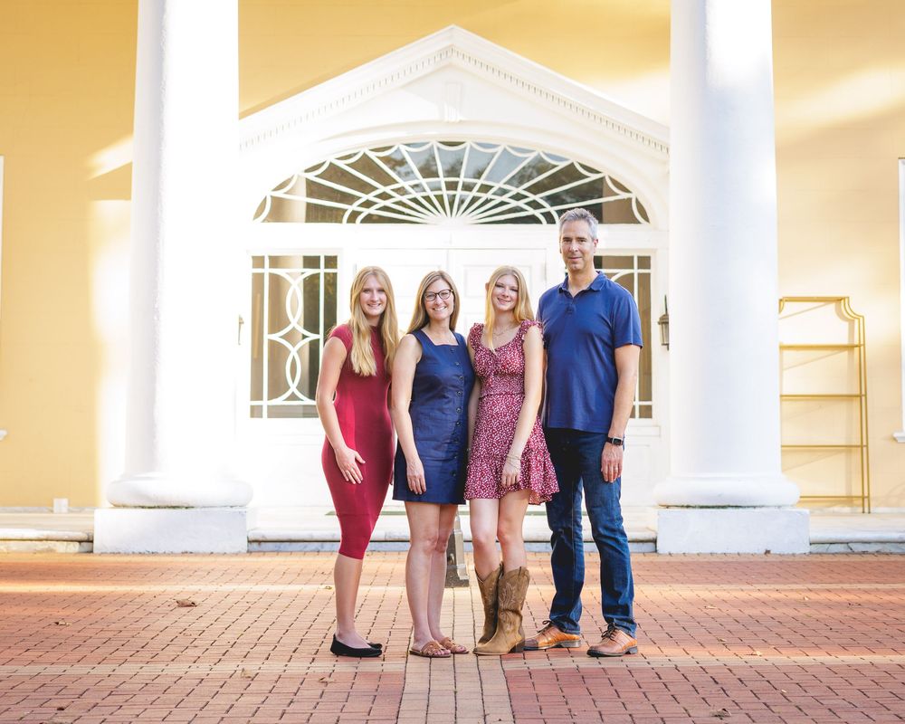 Family of four smiling together outside a yellow building with white arched entrance on a brick walkway.