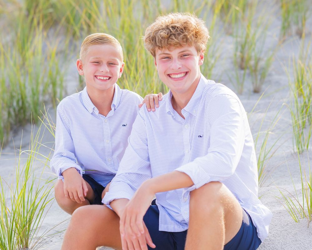 Two smiling teenage boys in matching blue shirts pose together on sandy beach dunes with sea grass.