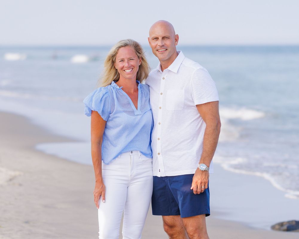Smiling couple posing together on a sunny beach, woman in blue shirt and white jeans, man in white shirt and navy shorts.