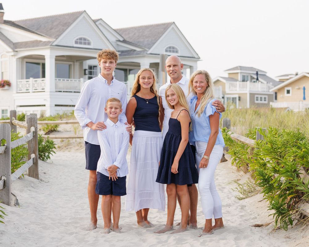 Family of seven posing together on a sandy beach path in front of a coastal home, wearing coordinated navy and white outfits.