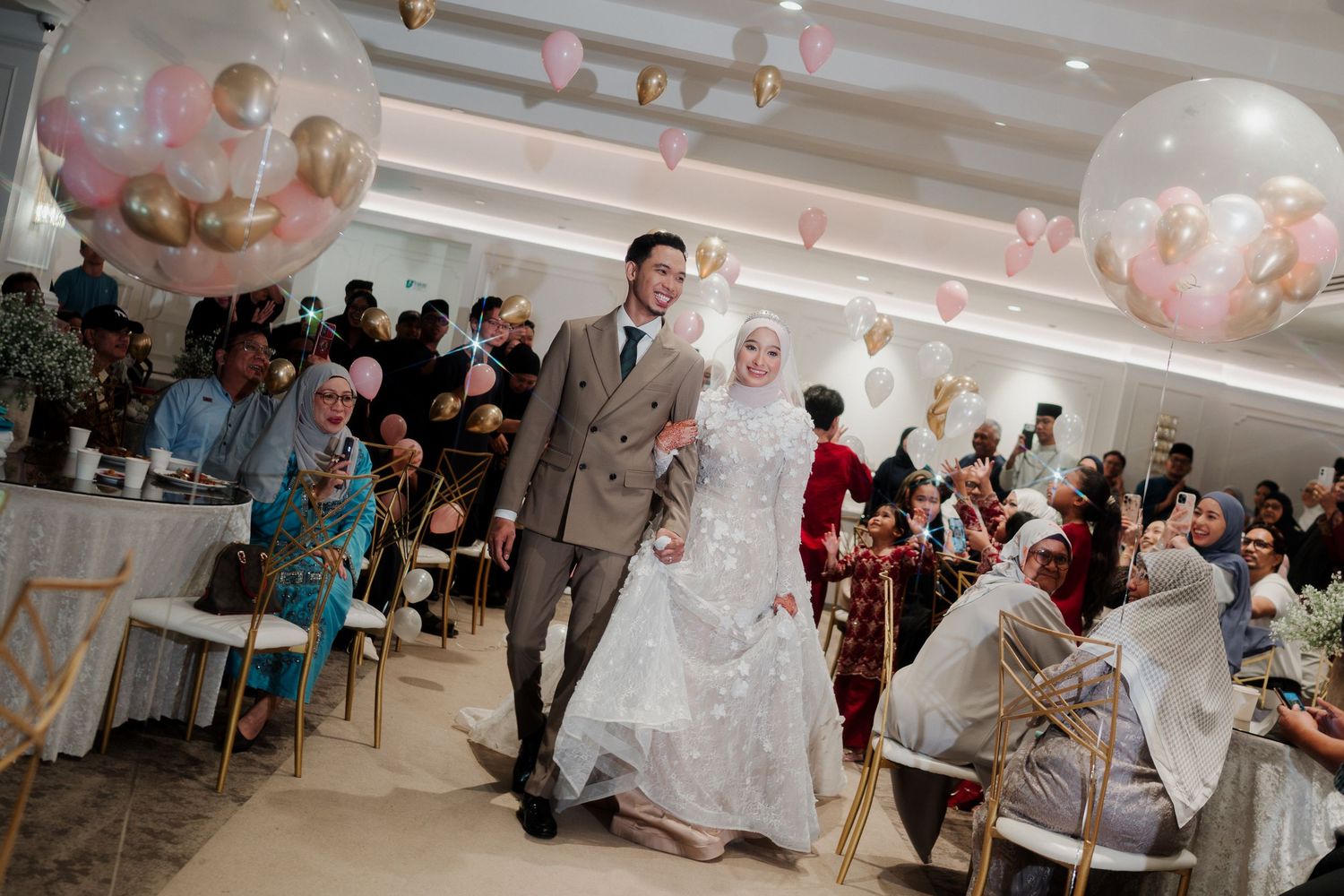 Muslim couple walks down the aisle at elegant wedding reception with pink balloons and guests celebrating.