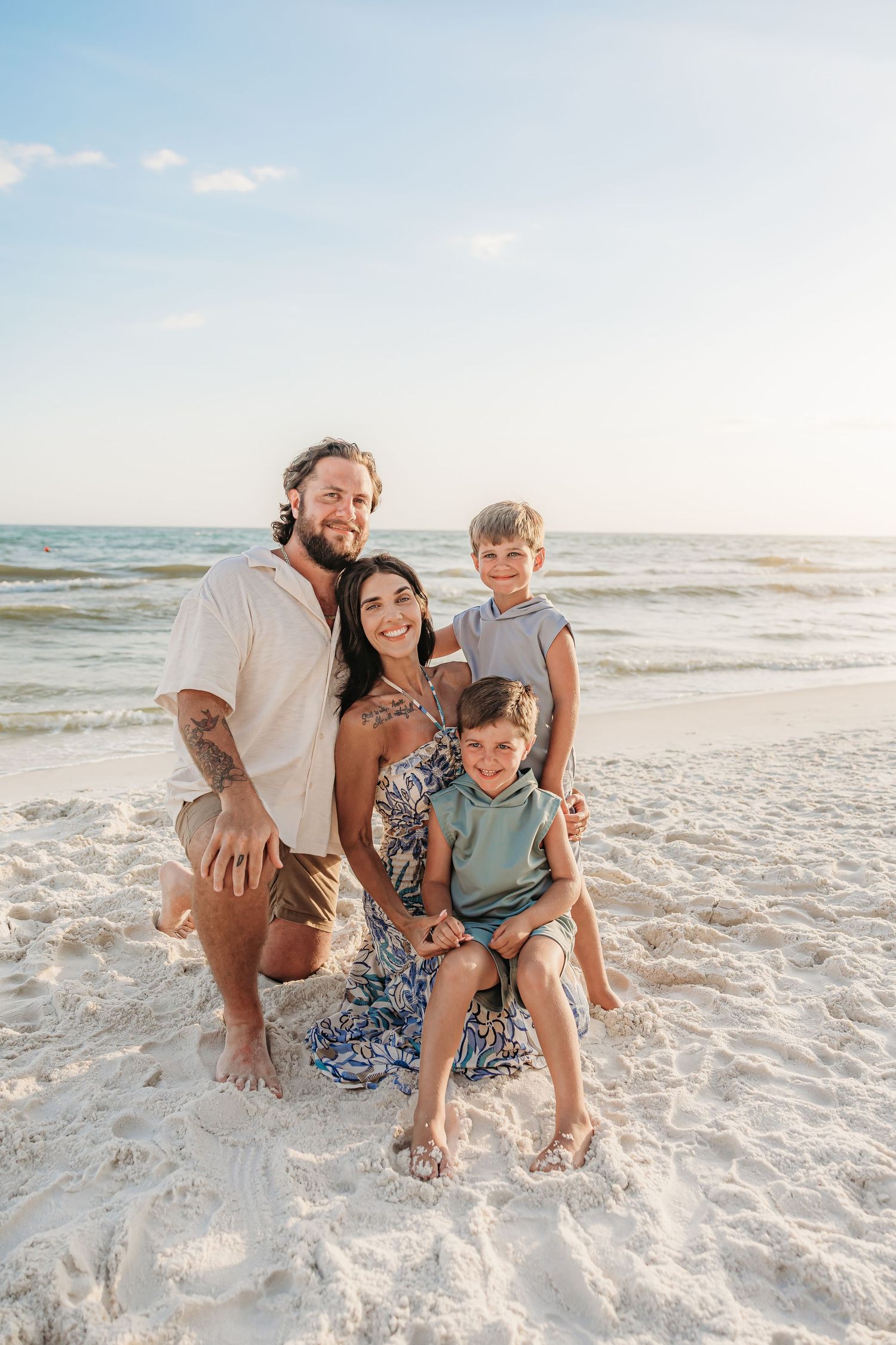 Family of five smiling together on a white sand beach near a pier on a sunny day.